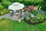 A table and chairs sit under a parasol in a lush garden